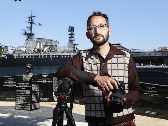 Evan Apodaca young man in glasses holding a camera standing in front of a warship