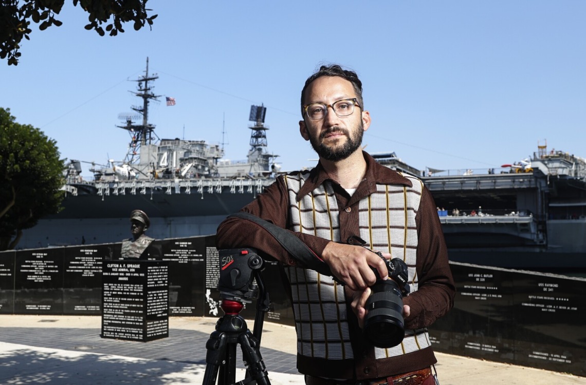 Evan Apodaca wearing a sweater, holding a camera, and standing in a park with a warship in the background