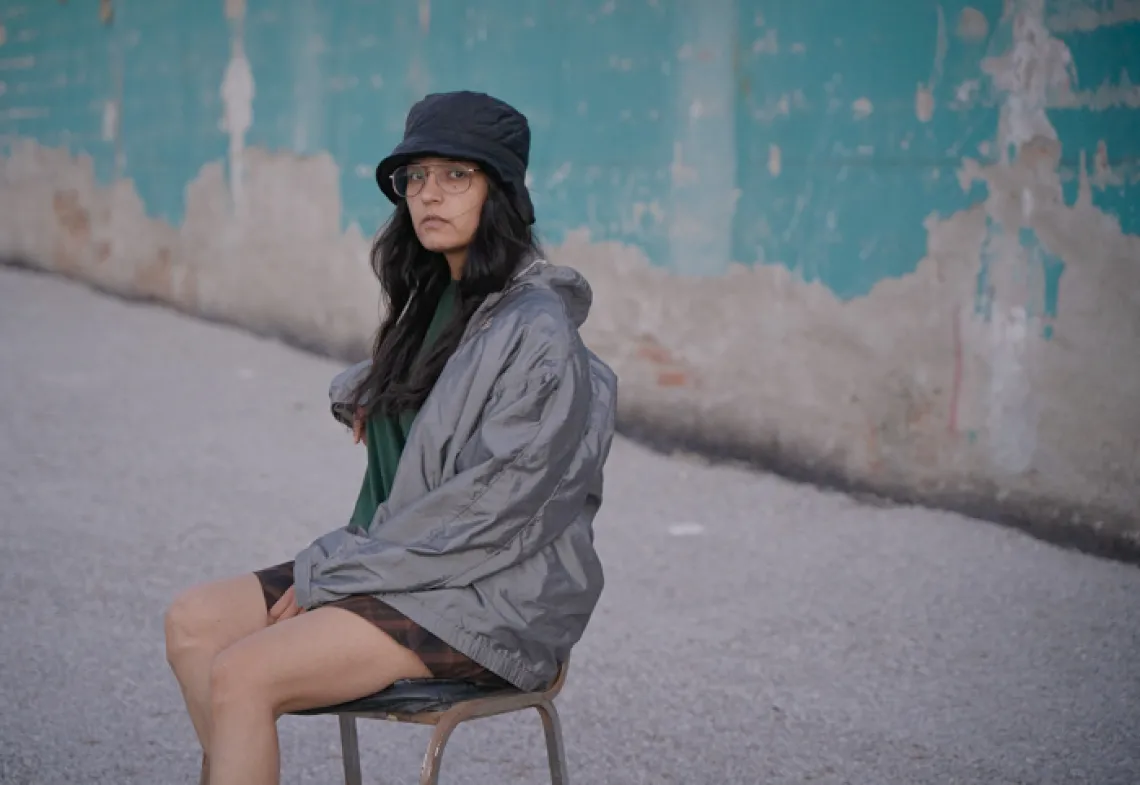 Alejandra seated in front of a blue wall wearing a dark hat