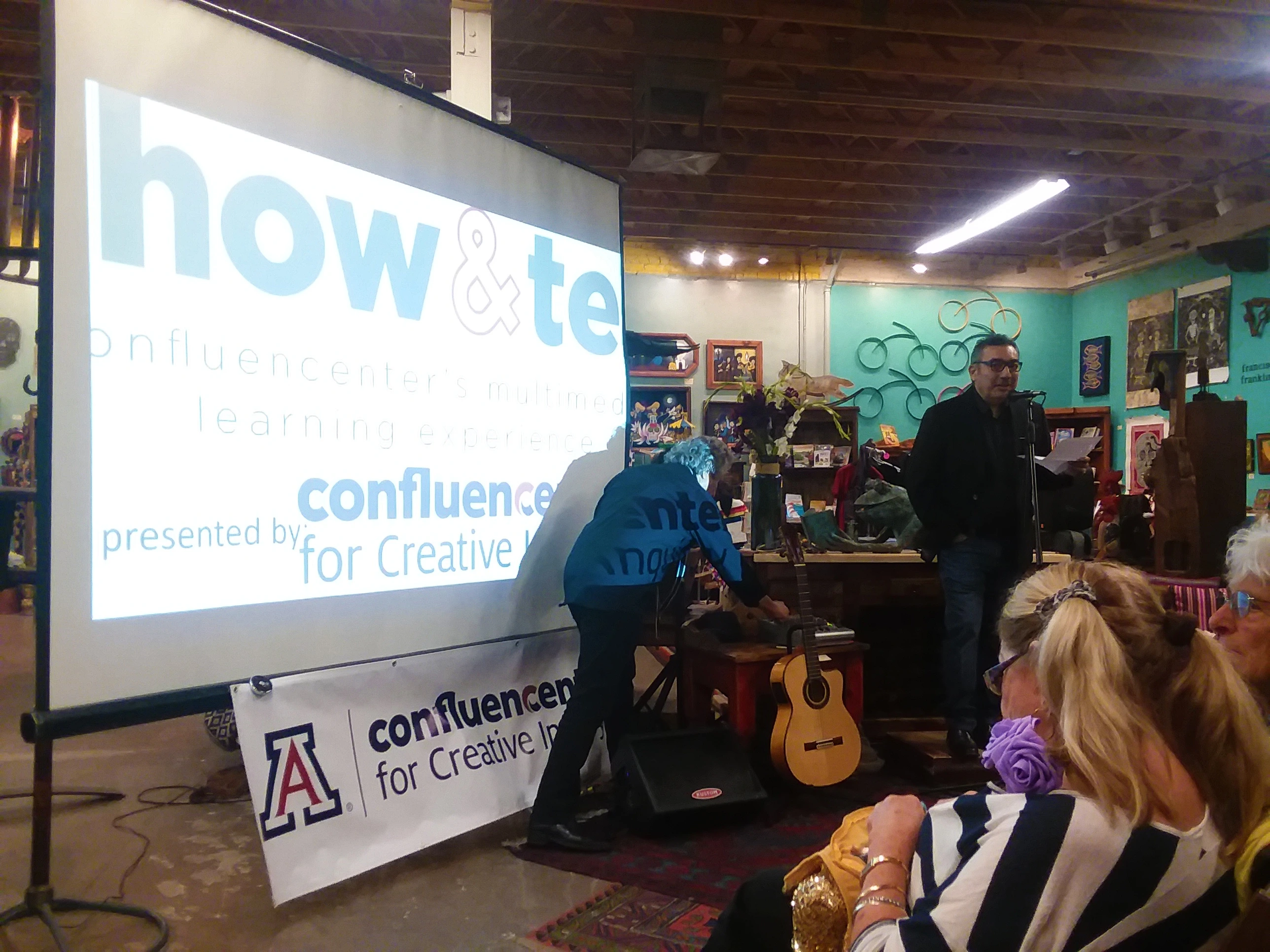 Show and Tell presentation in a room with teal walls and exposed wood beam ceiling. Dr. Javier Duran speaks at a microphone on the right while another man reaches for a guitar in the center. A woman with blonde pigtails and a purple rose lapel pin sits in the foreground. A large projector displays the presentation.