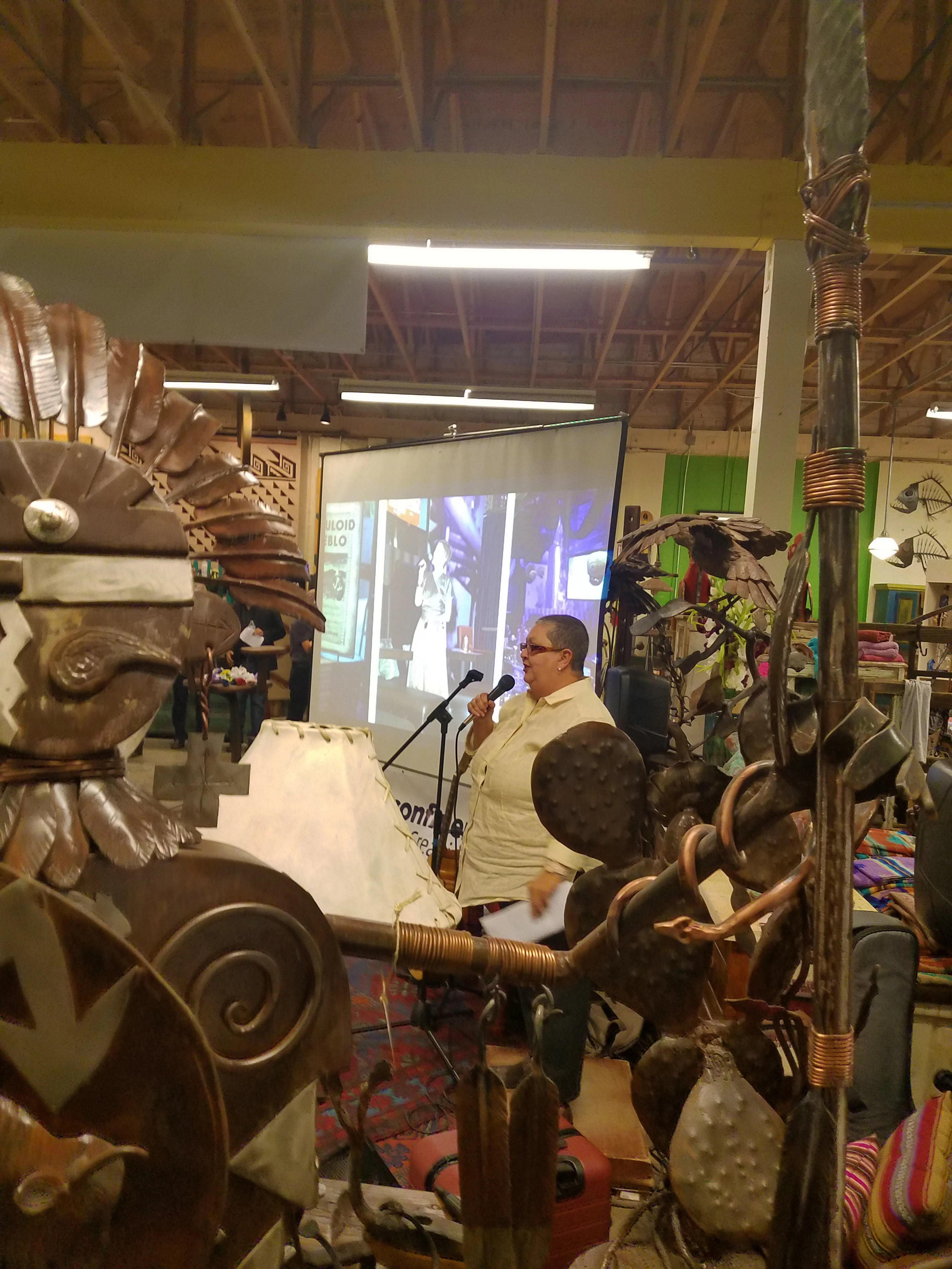 Show and Tell presentation in a room with teal walls and exposed wood beam ceiling. A presenter speaks at a microphone. A large projector displays the presentation.