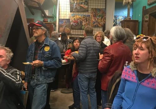 People gathering in a shared space waiting for a discussion to start with a focus on an older man in a jean jacket and red hat. There is also a women in a blue coat with a purple lapel.