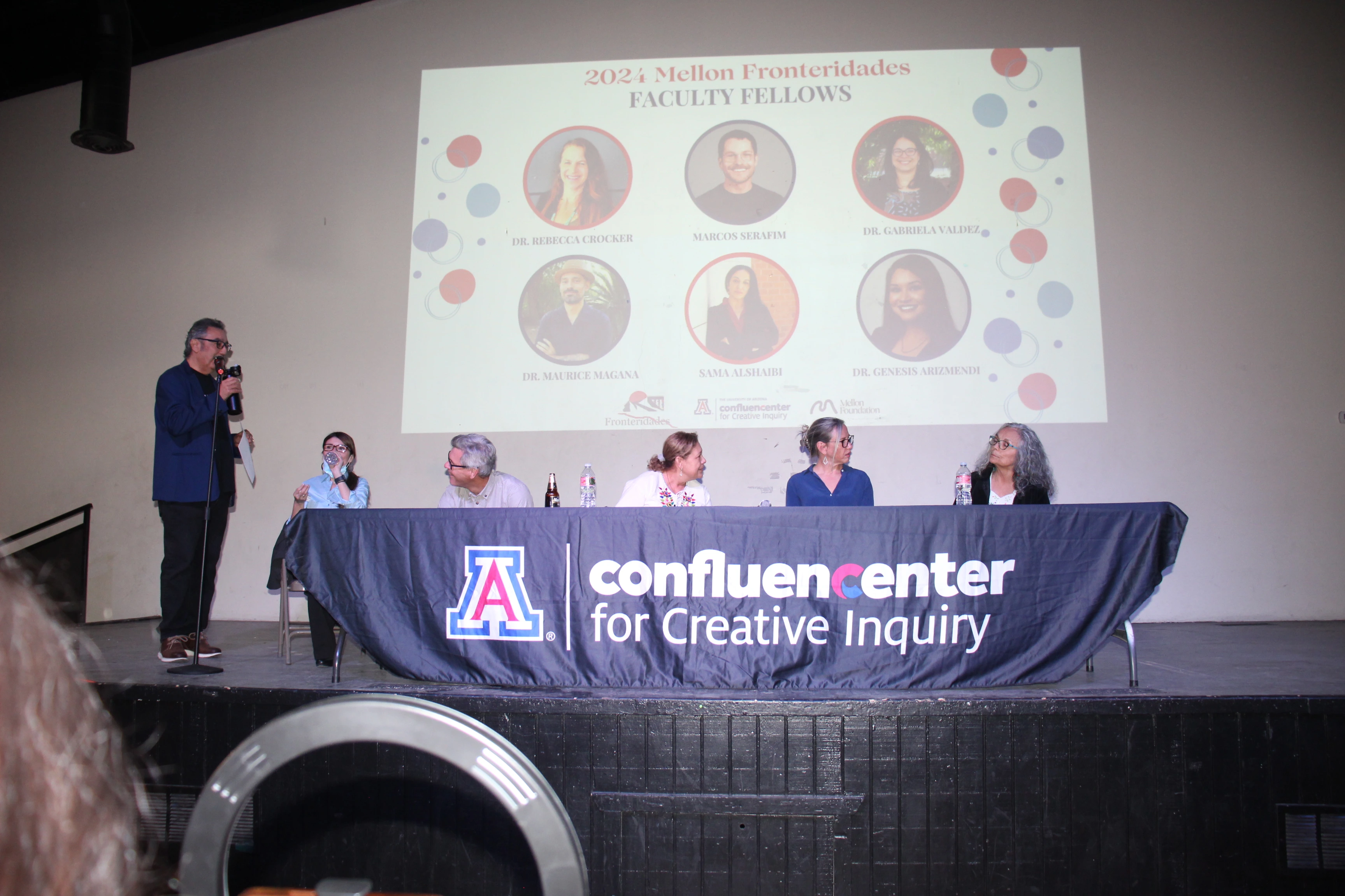Man with microphone talking to a panel on stage