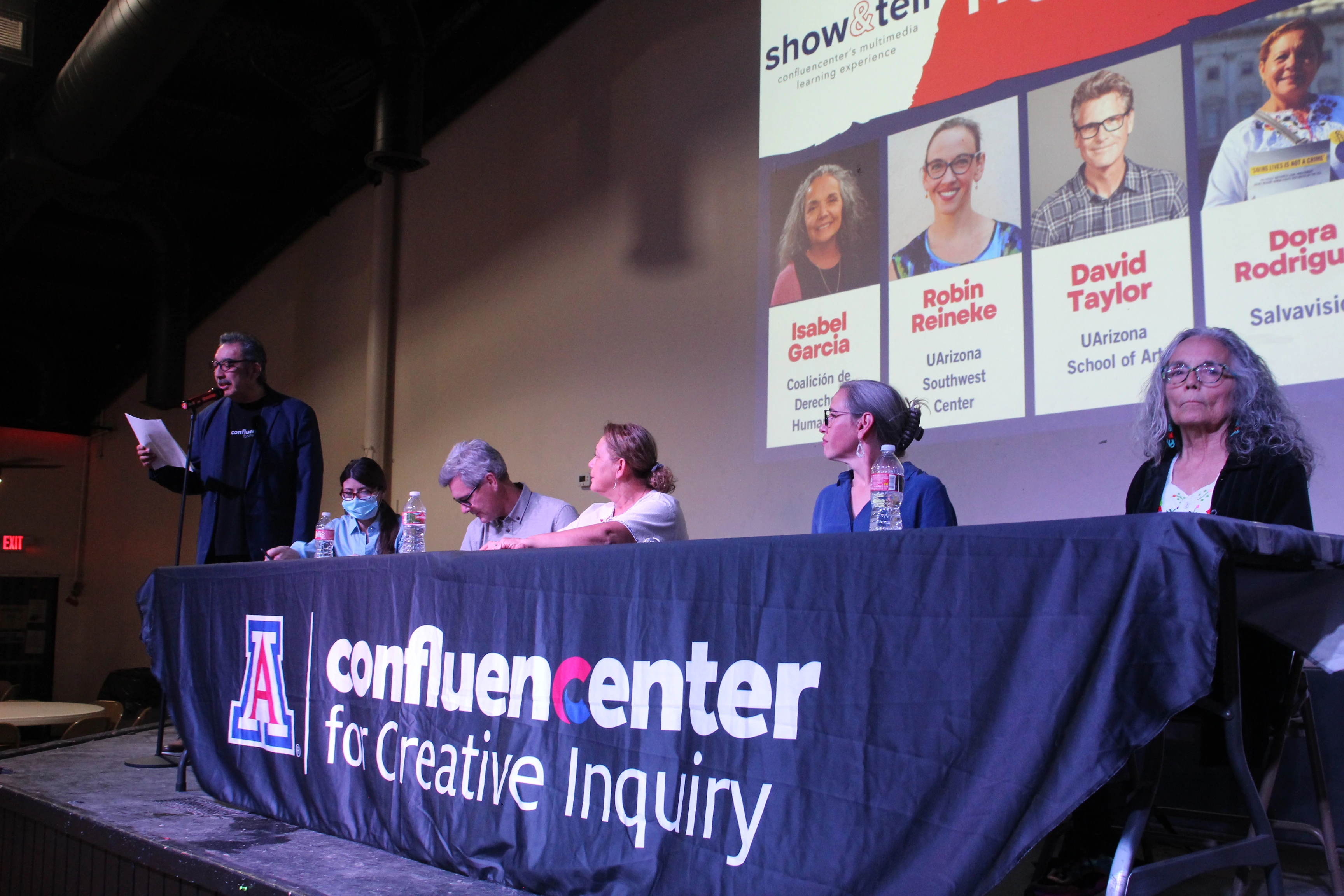 Man with microphone reading to a crowd on stage, with panel.