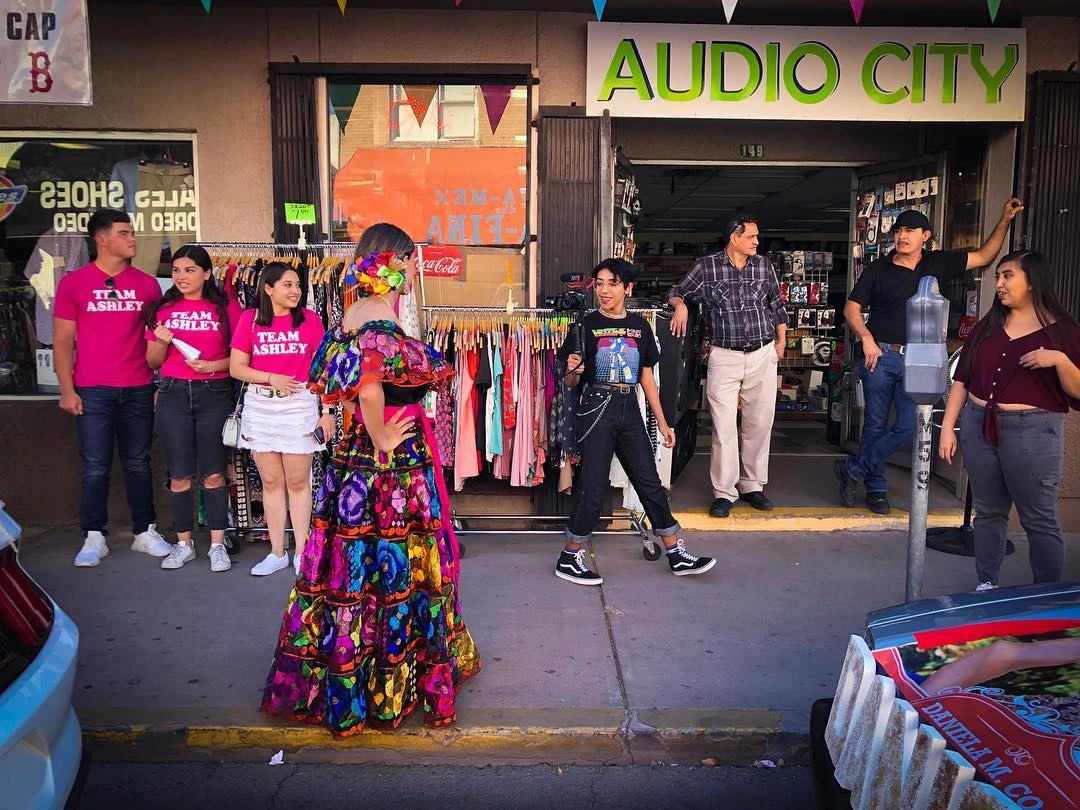 A women in a beautiful traditional Mexican style dress walking along a sidewalk. One old man is sitting in a chair, another man leaning in a doorway of a storefront along the walk. There is a small group of people with pink shirts as behind the women in the dress along the wall.