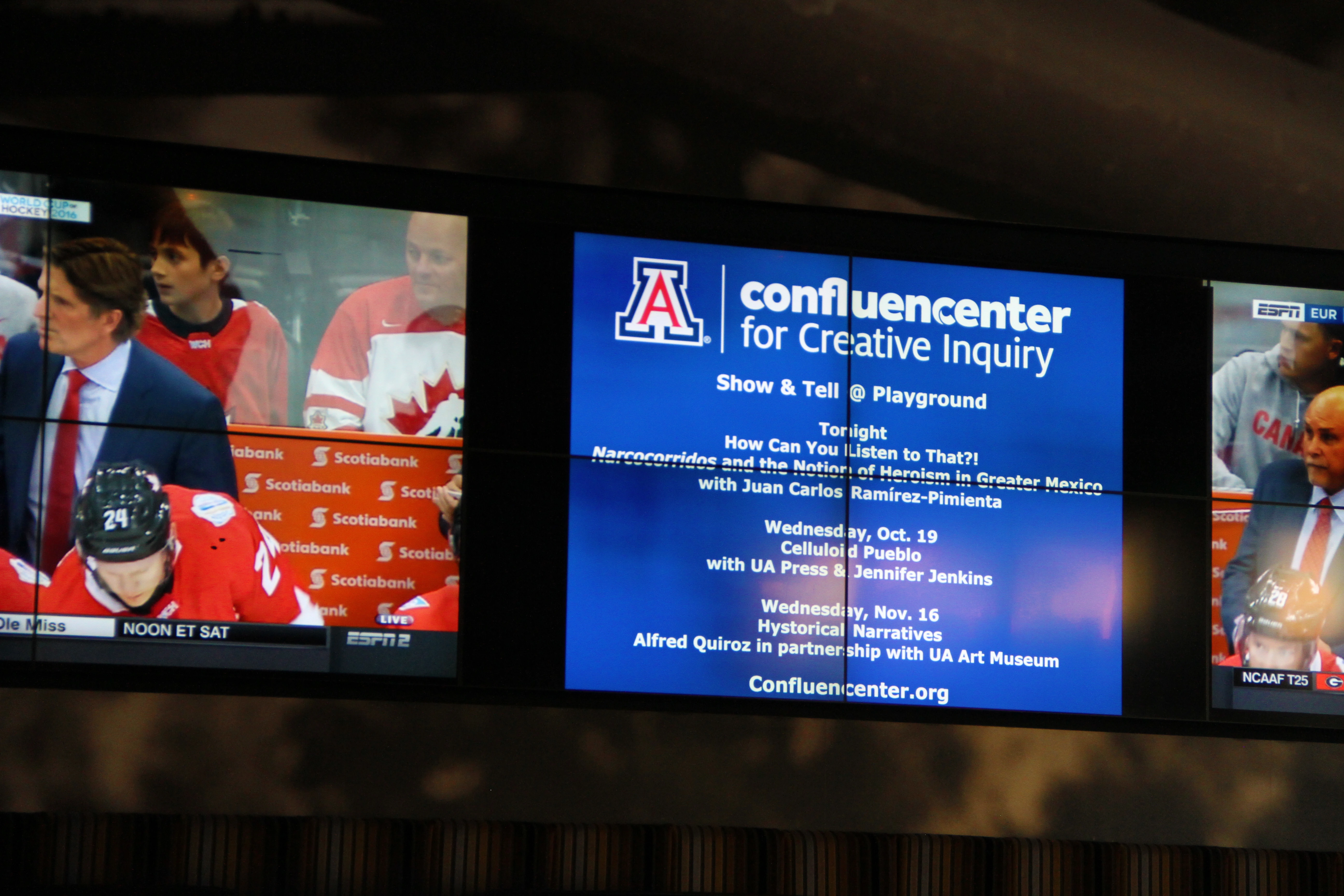 Advertisement for Confluencenter displayed on arena video board during professional hockey game. The venue features three large video displays, each made of four smaller screens. The center display shows the Confluencenter ad, while the left display shows a coach speaking to players at the bench.